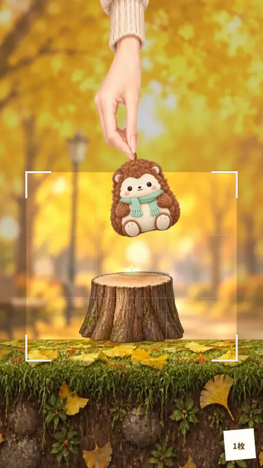 スマホの縦画面。ぼけたイチョウ並木の公園を背景に、上からのびた手がハリネズミのぬいぐるみをつまんで、落ち葉の芝生の上の切り株へ運んでいる。まわりには写真の枠のしるし